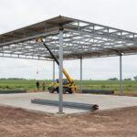 Galpón agropecuario de estructura metálica en construcción sobre platea de hormigón, con manipulador telescópico y perfiles en el campo